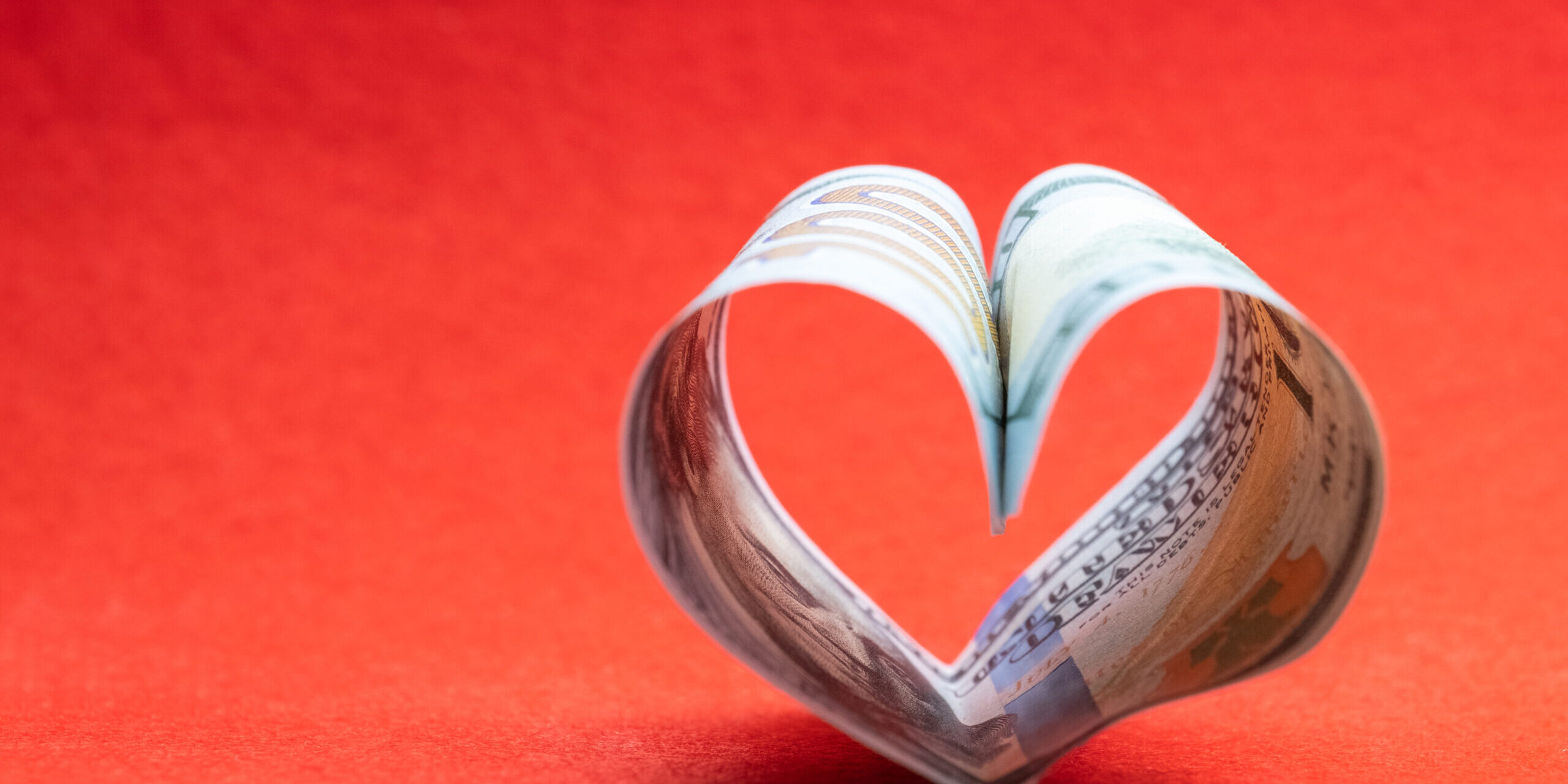 US 100 dollar bill in the shape of a heart on a red background. Copy space. Concept of money, love and a gift for Valentine’s Day.