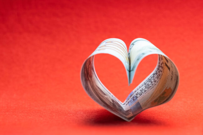 US 100 dollar bill in the shape of a heart on a red background. Copy space. Concept of money, love and a gift for Valentine’s Day.